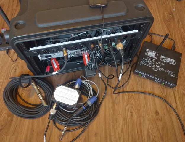 A rear view of the system with connected accessories including an XLR cable, a signal attenuator, a speakON cable, a transformer/splitter box, an assistive listening transmitter, and a cell phone